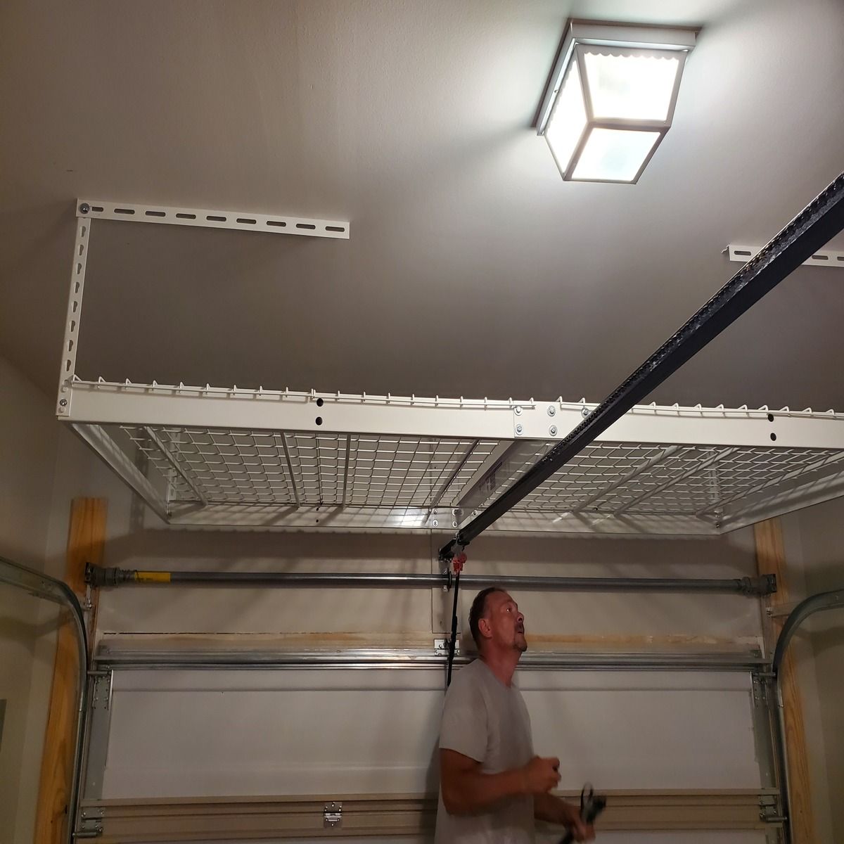 Man in garage looks up at overhead storage shelf. The garage door is below and the ceiling has a light fixture.