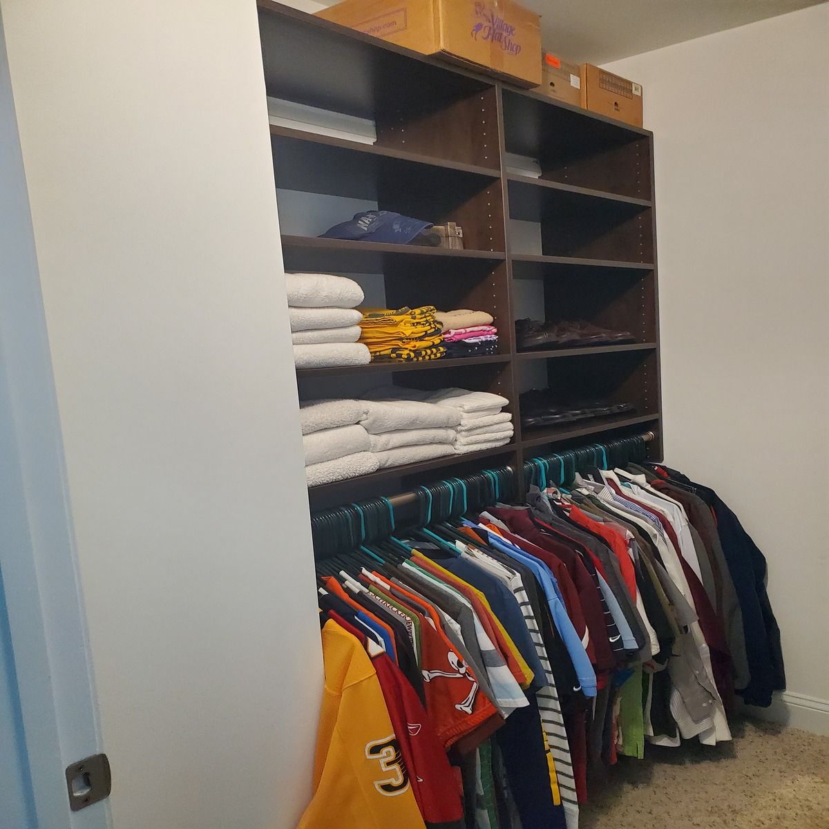 Closet with dark wood shelves and hanging clothes. Includes folded towels, shirts, and cardboard boxes on top shelf.
