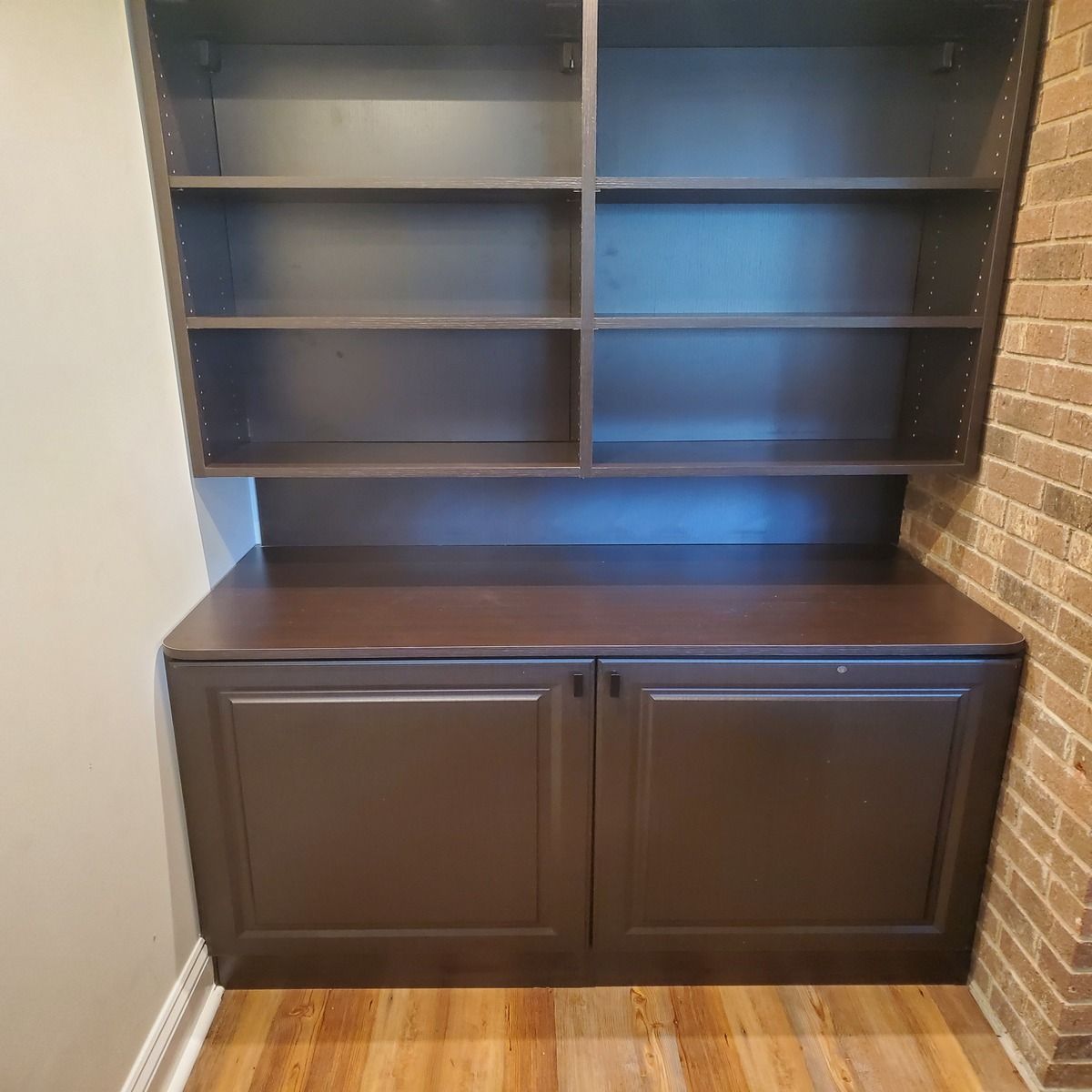 Built-in dark brown cabinet with open shelves above and two cabinet doors below. Set in a corner between brick and painted walls.
