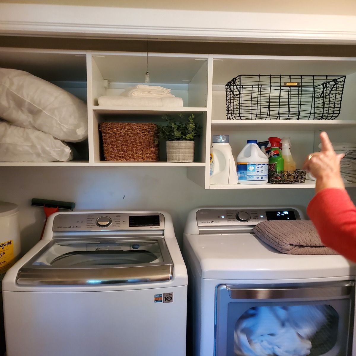 Laundry room with white appliances and overhead shelving. A hand points at a wire basket on a shelf filled with cleaning supplies and baskets.