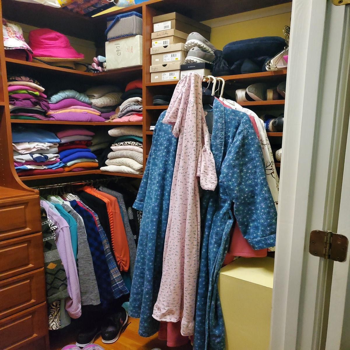 A cluttered walk-in closet with clothes, shoes, and boxes. A blue robe and pink dress hang on a rack.