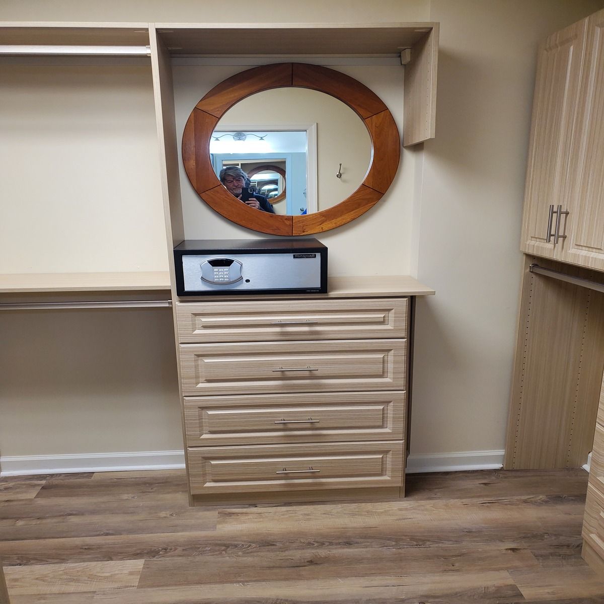 A closet interior features a light wood dresser, a safe on top, a round mirror, and wood flooring. A small glimpse of a person is reflected in the mirror.