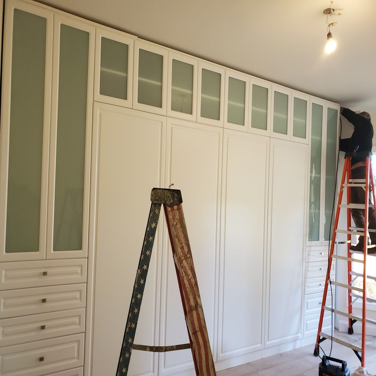 A person on a ladder installing a custom white built-in closet with frosted glass panels.