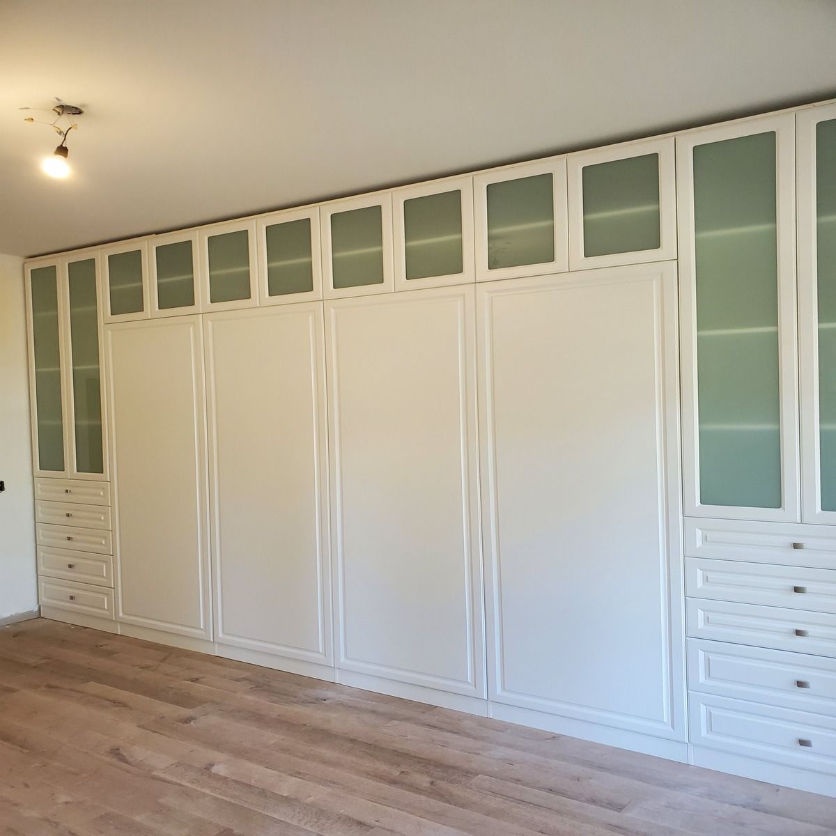 Built-in white closet with multiple doors and glass-paneled upper cabinets. It's in a room with wood flooring.