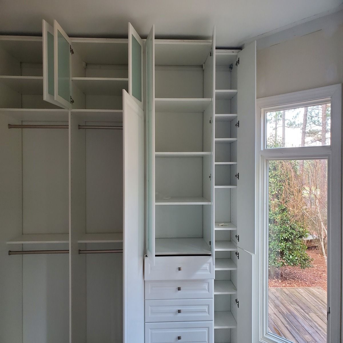 White built-in closet with open doors revealing shelves and drawers. A window with a view of outdoors is on the right.