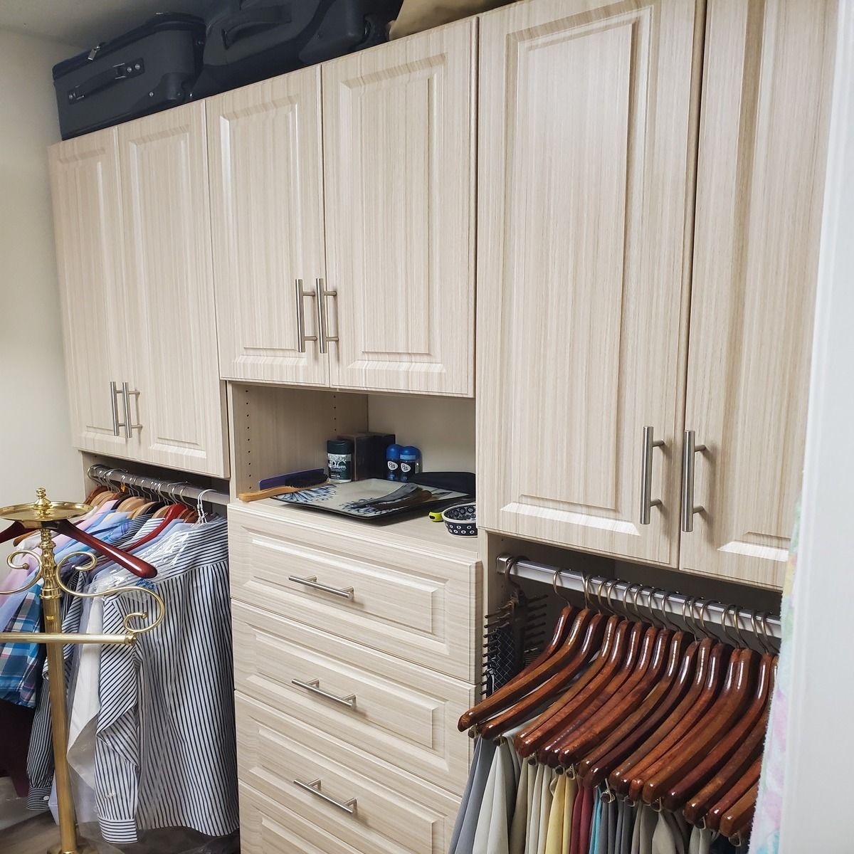 Closet with light wood cabinets, drawers, and hanging rods. Clothes and suit bags fill the rods.