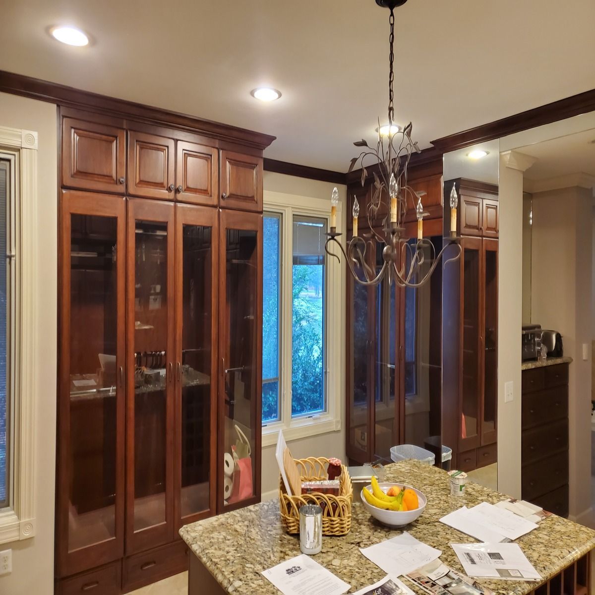 A kitchen with dark wood cabinets and a granite countertop island. A chandelier hangs above the island with fruit and papers.