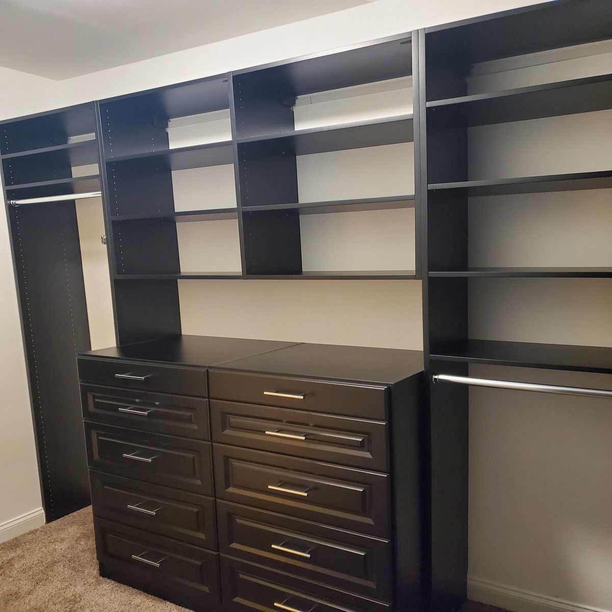 Black closet organizer with shelves, drawers, and hanging rods, in a room with tan carpet and white walls.