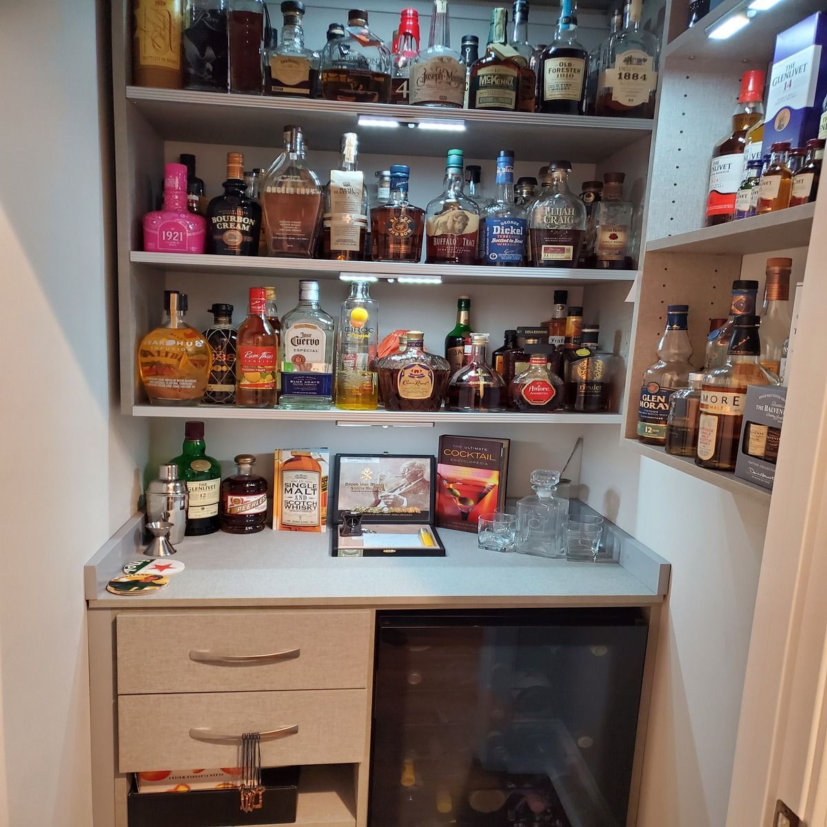 A built-in home bar with multiple shelves filled with liquor bottles, a mini-fridge, and a small countertop area.