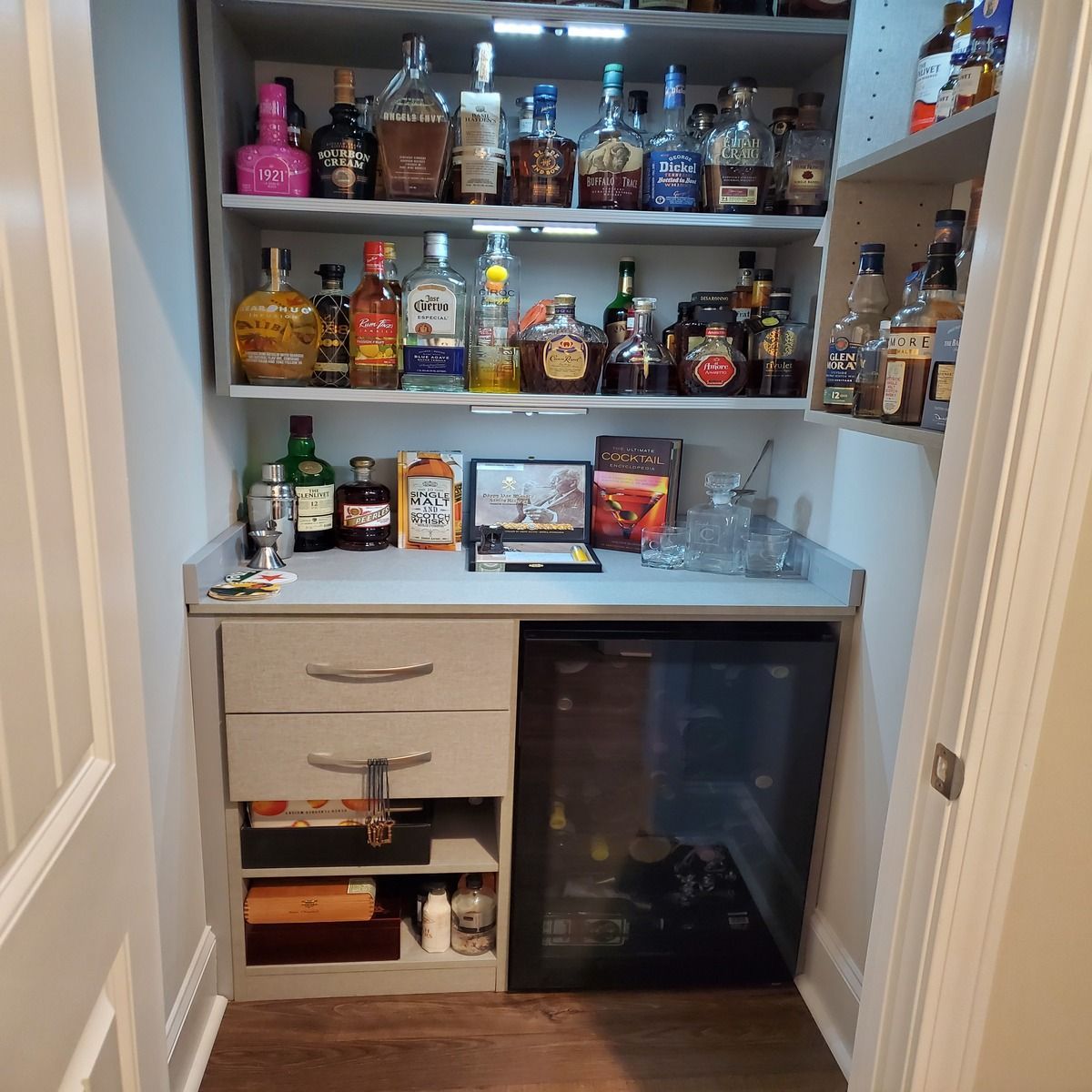 A small home bar built into a closet. Grey cabinets with shelves holding liquor bottles, a small countertop, and a mini-fridge.