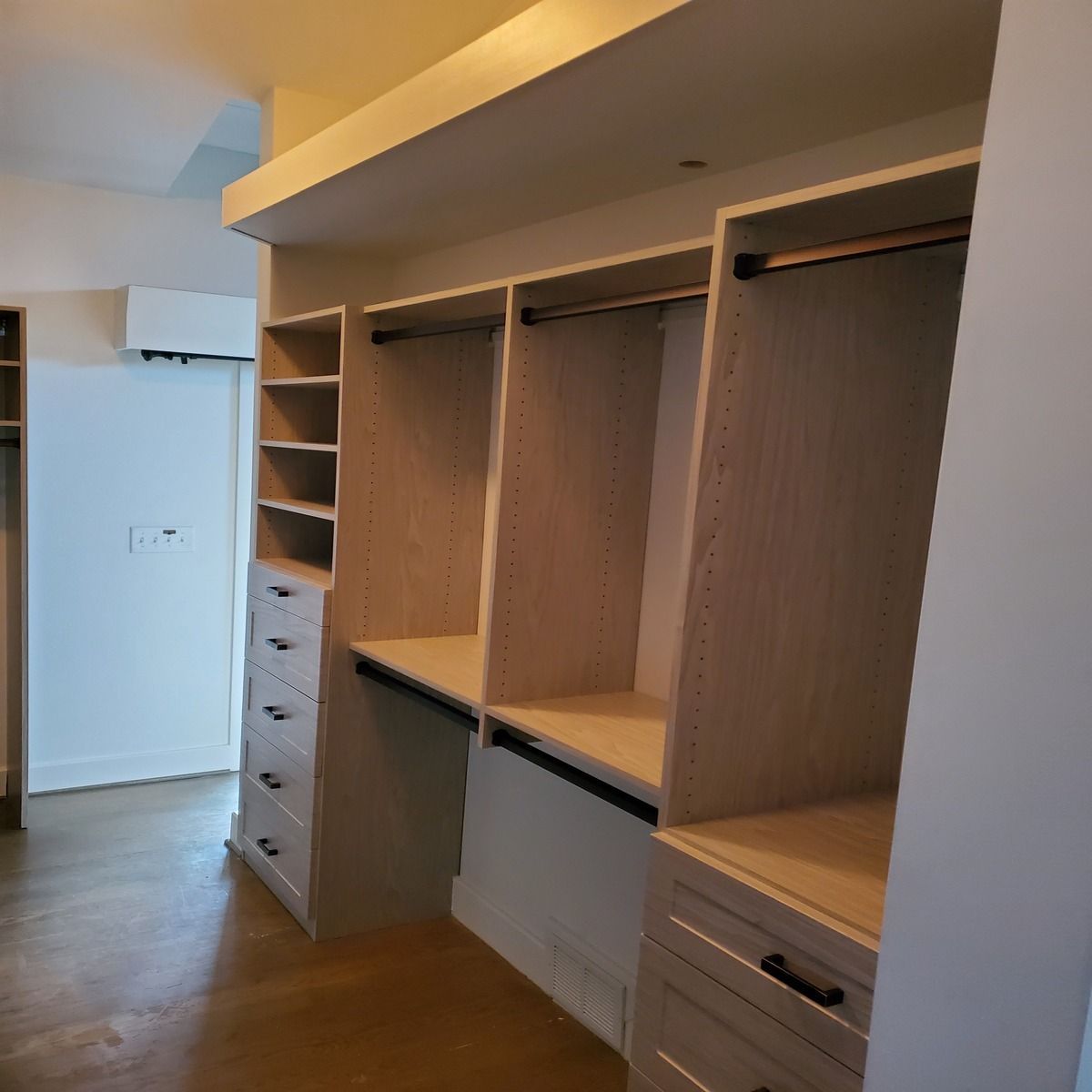 Built-in closet with shelves, drawers, and hanging rods. Beige wood and black hardware are in a white-walled room.