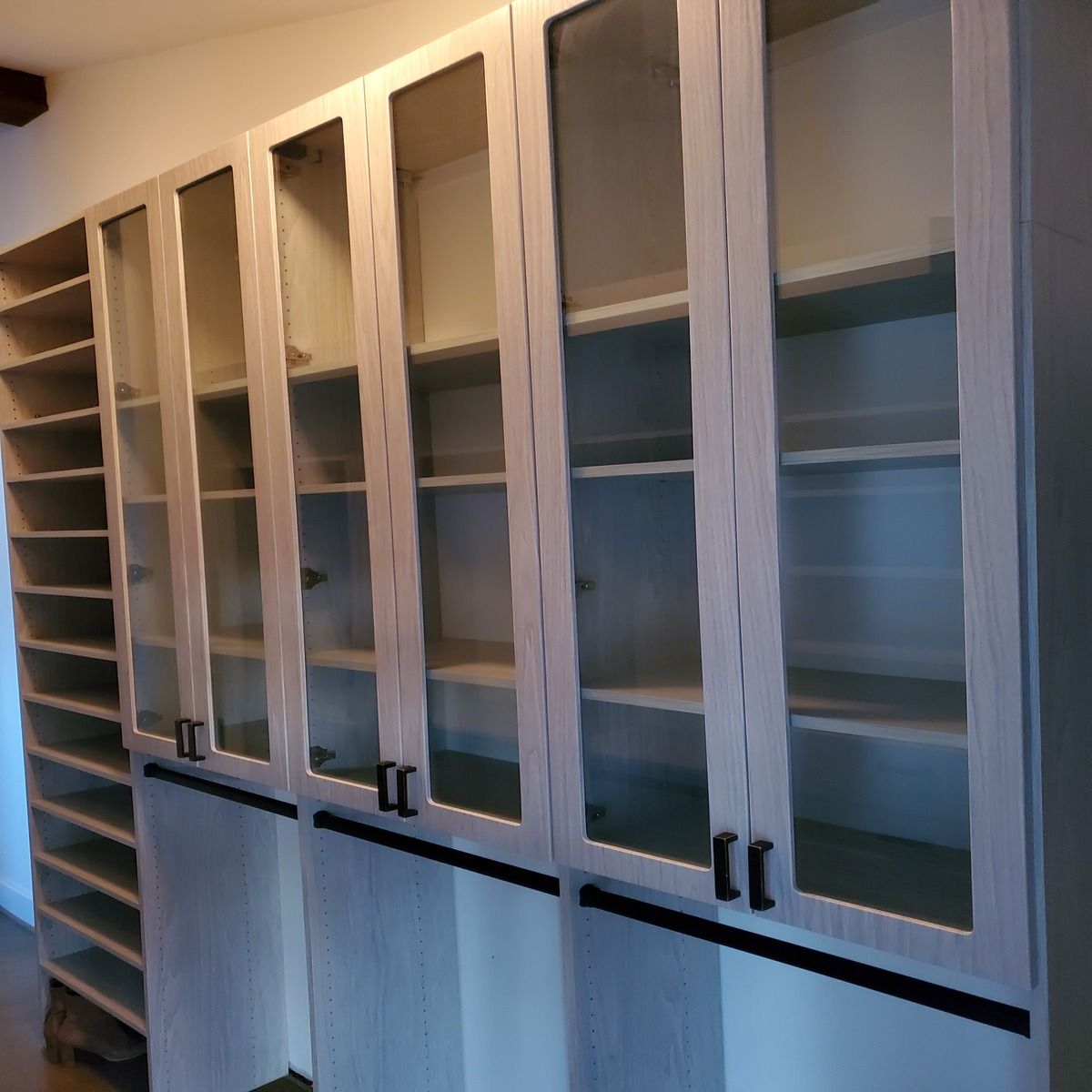 Built-in cabinets with glass doors and black hardware mounted on a wall. To the left is a tall open shelving unit.