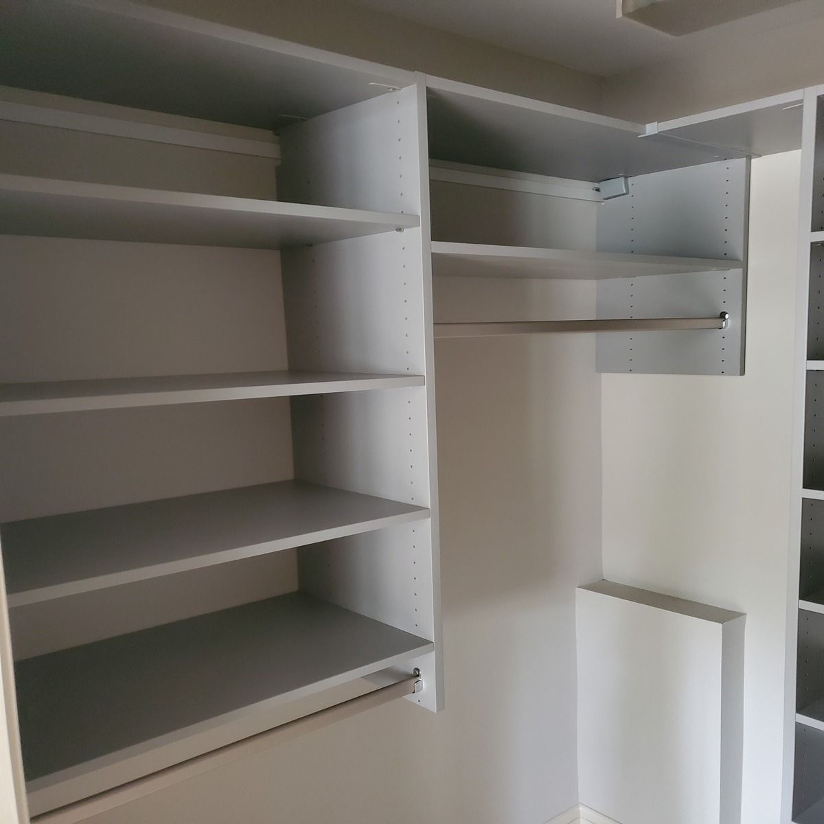 A built-in closet with light gray shelves and a clothes rod. Empty, the corner closet is located in a room with white walls.