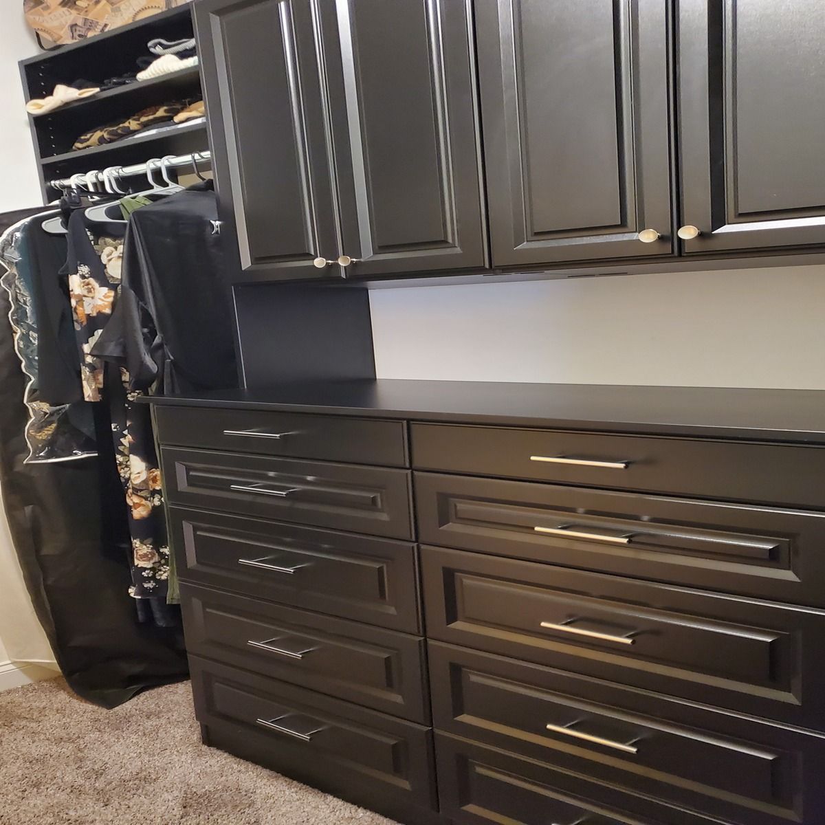 Black walk-in closet with cabinets and drawers. Clothes hang on a rack and on shelves above.