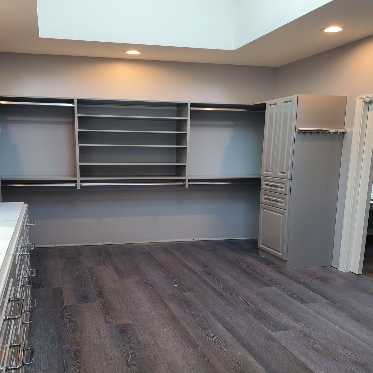 Empty gray walk-in closet with shelves, rods, and flooring. Includes a cabinet and natural light from a skylight.