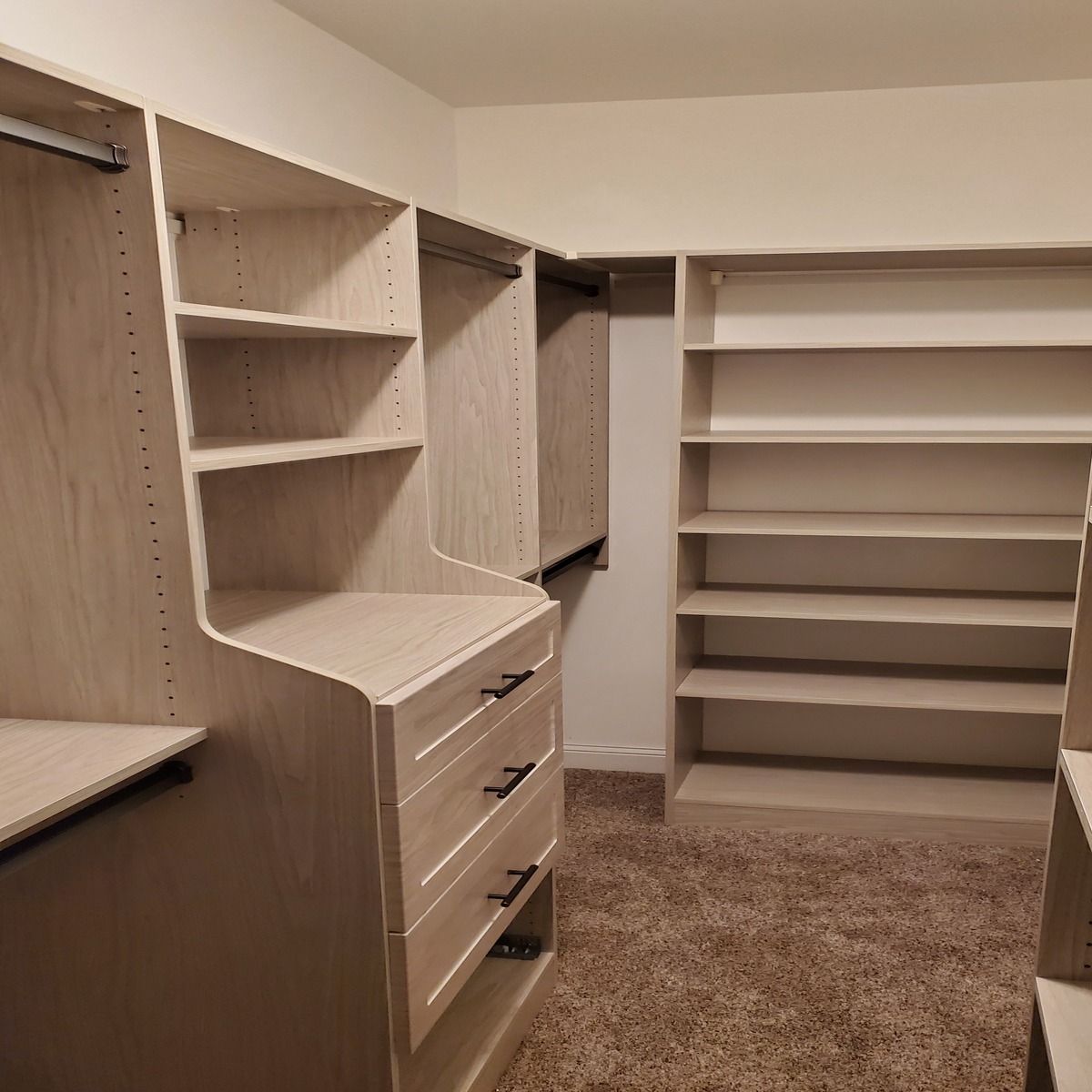 A walk-in closet with light wood shelving and drawers. The closet has a brown carpet and a white ceiling.