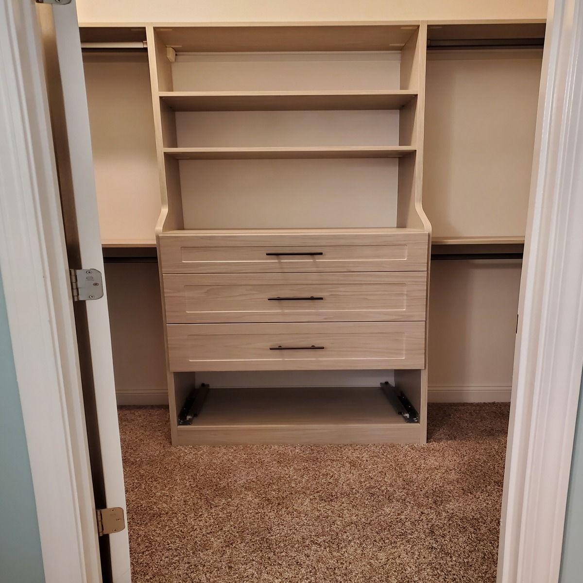 A light-colored custom closet organizer with shelves and drawers, centered in a closet with tan carpet and hanging rods.
