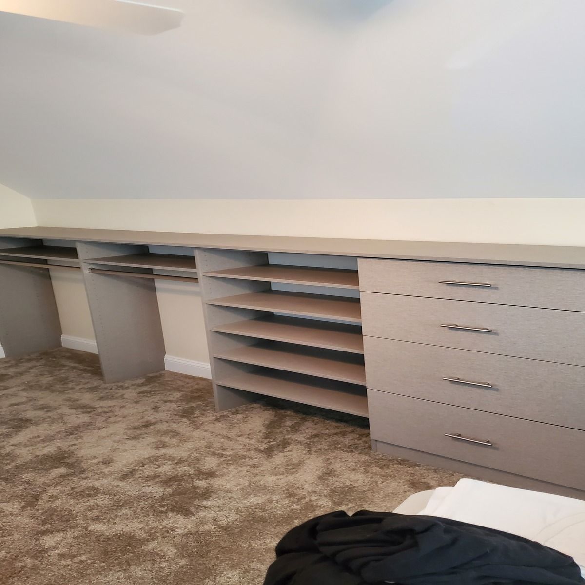 Built-in closet with shelves and drawers against a white wall and beige carpet. Light grey cabinetry is installed under a slanted ceiling.