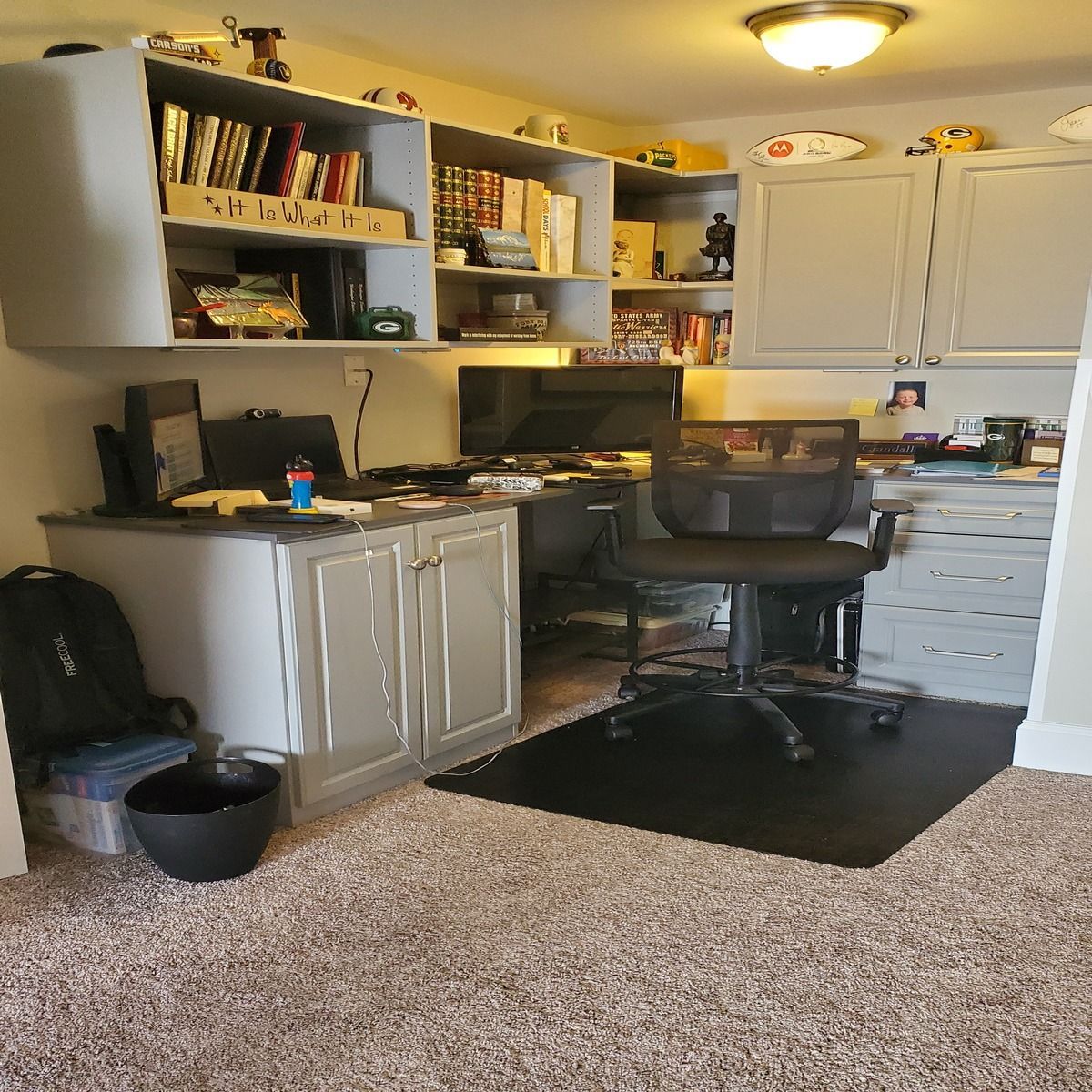 Home office with built-in desk, shelves, and cabinets. A black office chair sits on a black mat. Books and decorative items fill the shelves.