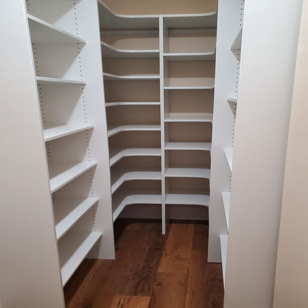 White built-in pantry with angled shelves, hardwood floor, and curved corner shelves; neutral wall color.