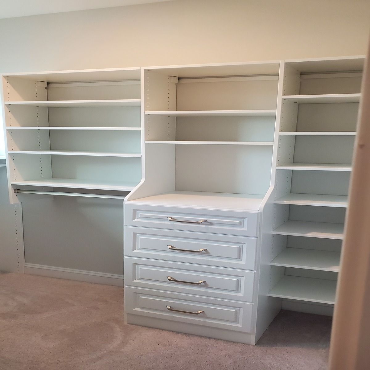 Built-in white closet with shelves, drawers, and a hanging rod, set against a light gray wall and carpeted floor.