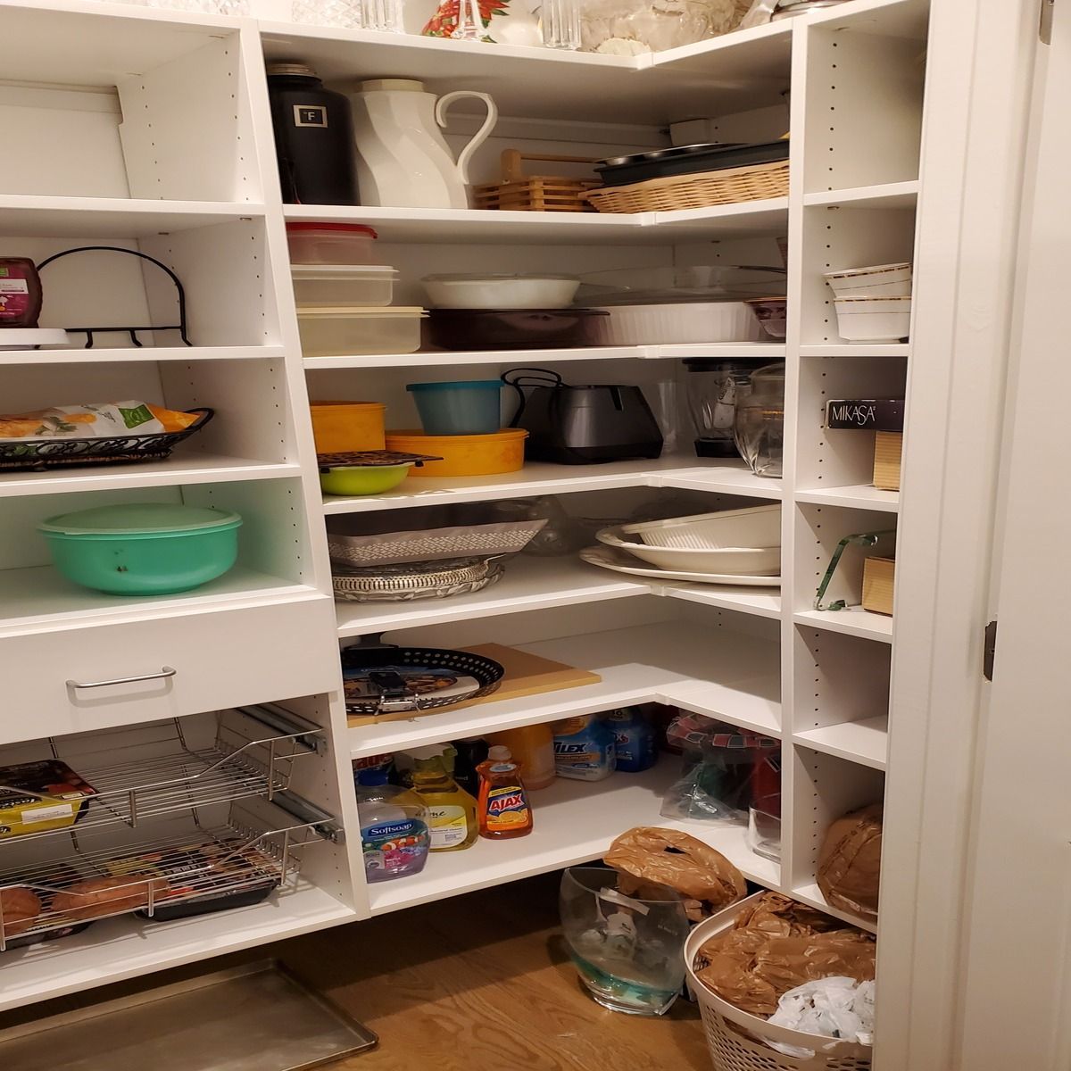 White pantry with shelves, partially stocked with various dishes, containers, and food items. Wooden floor.
