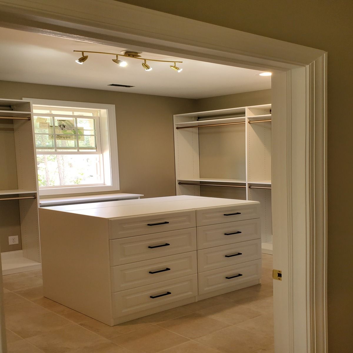 A walk-in closet with white shelving and a central island with drawers. Sunlight streams through a window.