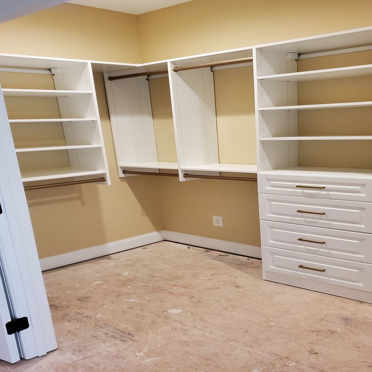 White closet organizer system in a corner of a room with beige walls and a concrete floor. Shelves and drawers are visible.
