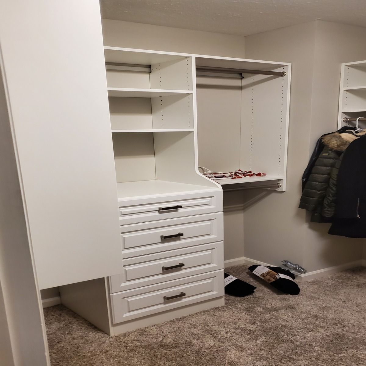 White walk-in closet with built-in shelves, drawers, and hanging rods. Carpeted floor, gray walls. Two pairs of shoes on floor.