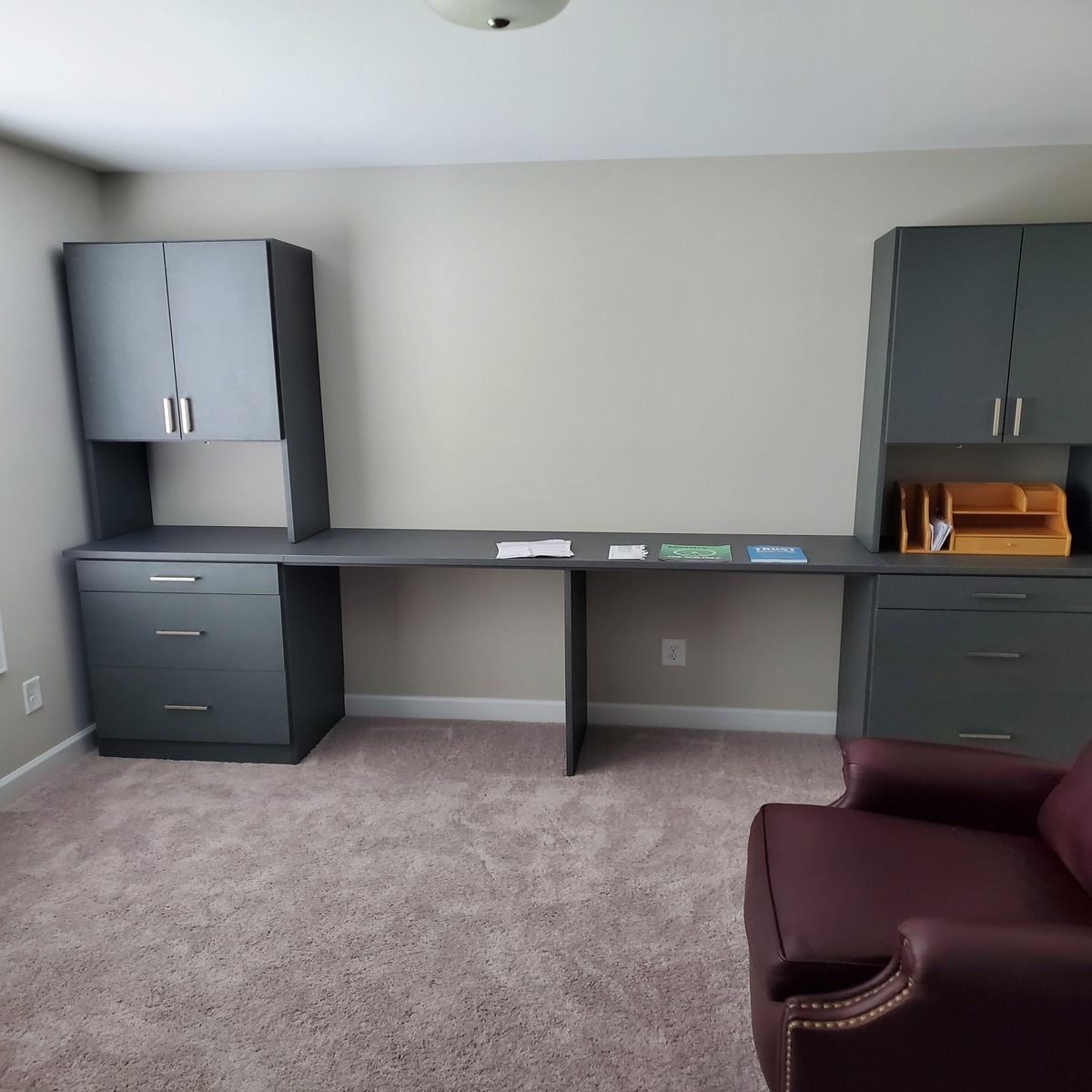 A home office with gray built-in cabinets and desk, tan walls, and burgundy armchair on a beige carpet.