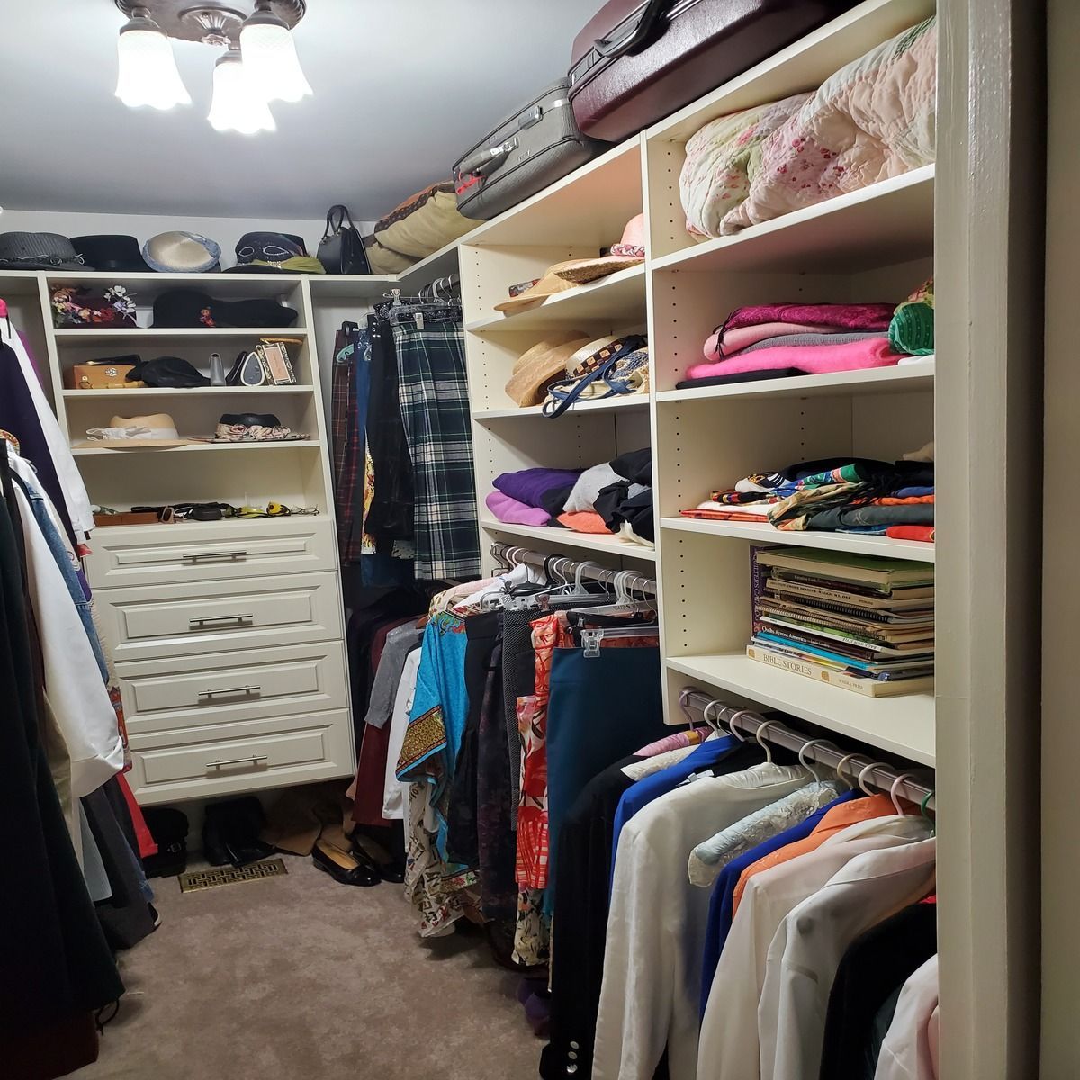 Walk-in closet with white shelving, filled with clothes, hats, shoes, and luggage. A light hangs from the ceiling, and the floor is carpeted.