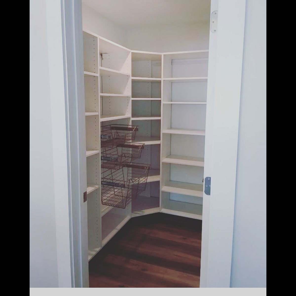 White pantry with built-in shelves and wire baskets, wooden floor. Open door reveals the storage space.