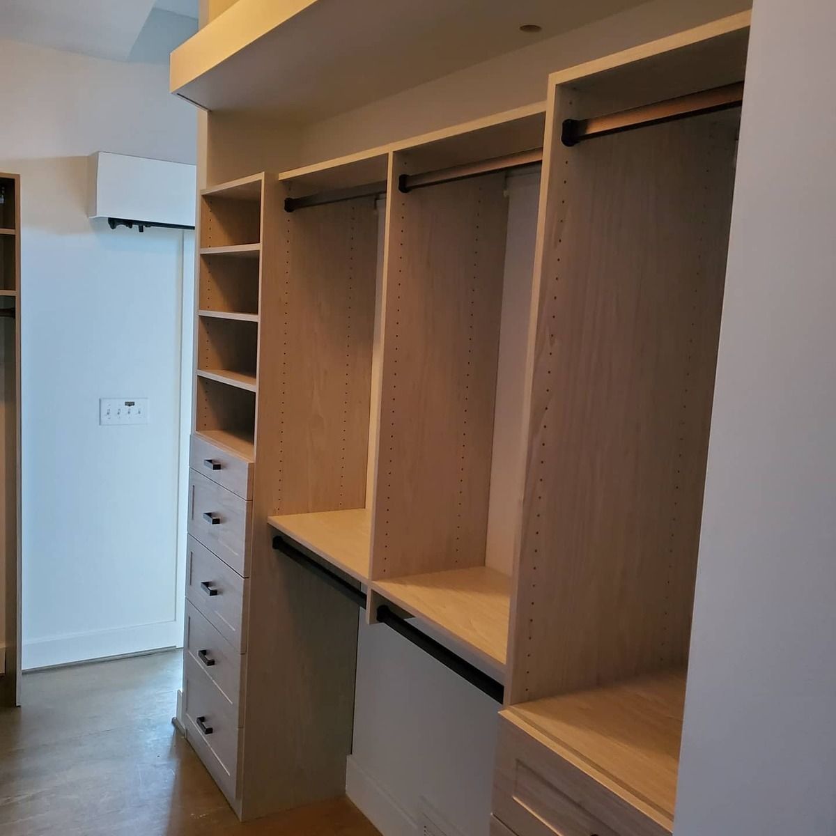 Custom-built closet with light wood shelving, drawers, and hanging rods. A neutral-colored room with a white wall and hardwood floor.