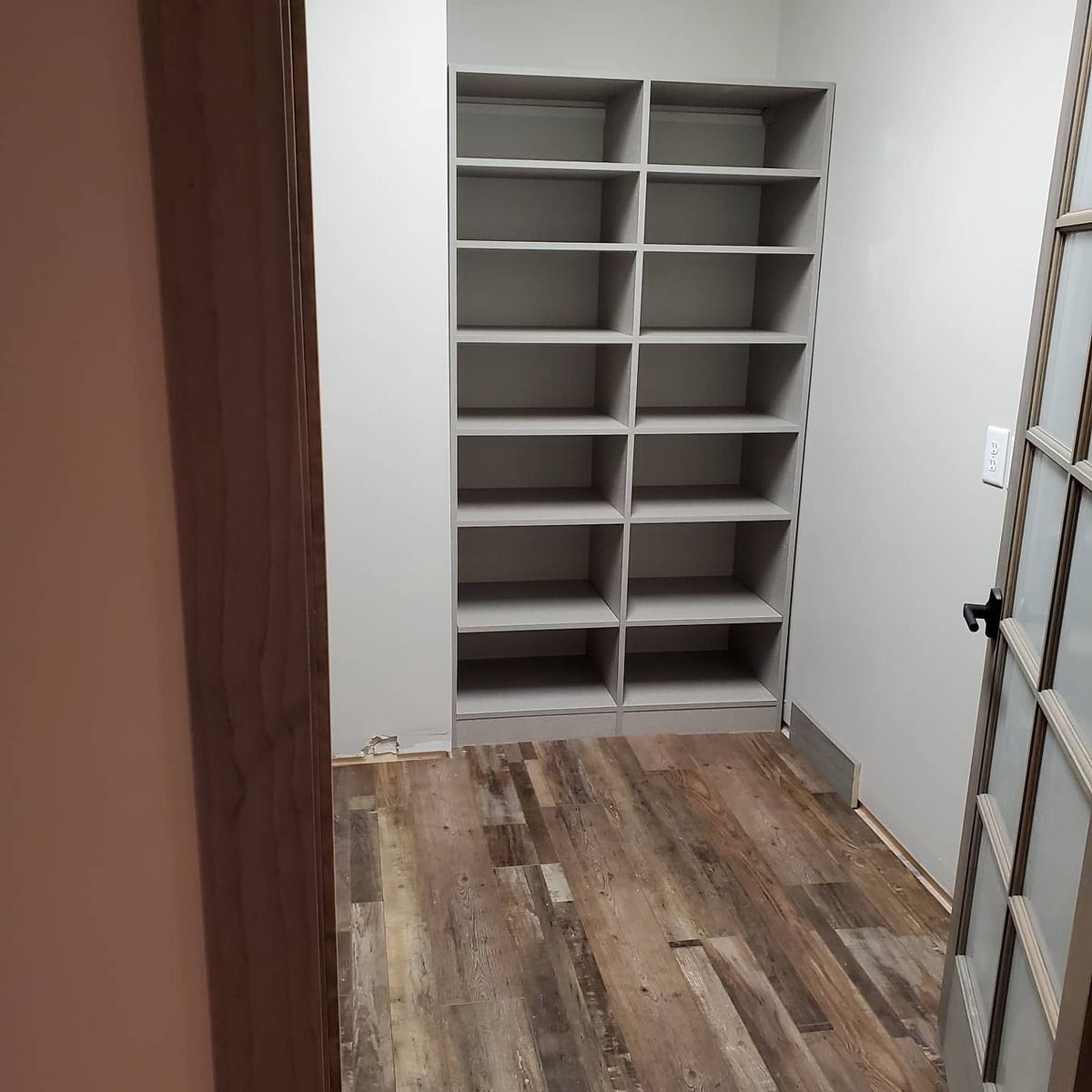 An empty built-in shelf unit in a room with wood-look flooring and a partially visible door. Walls are painted gray.
