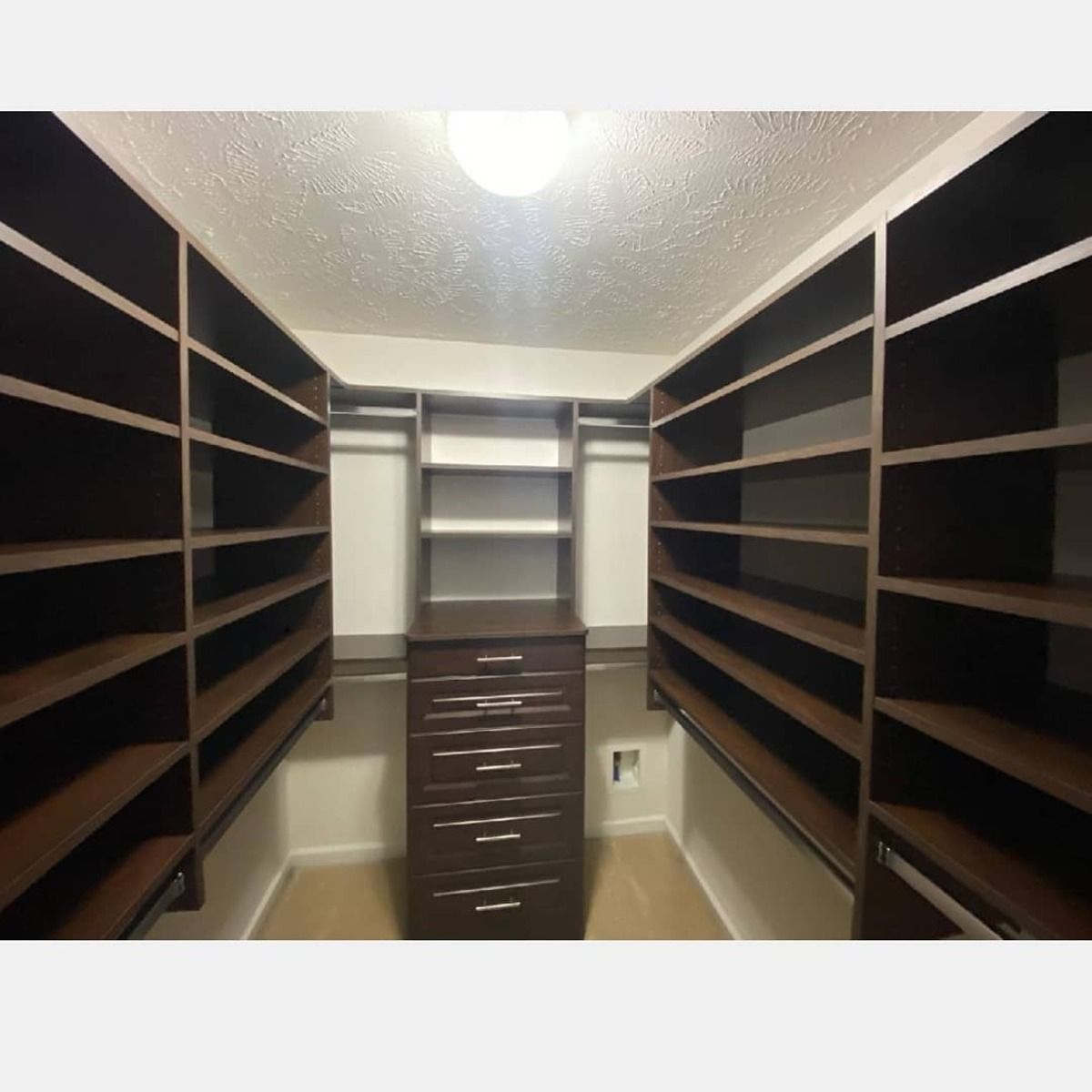 Empty walk-in closet with dark brown shelves and a central dresser. The walls are beige, and a light fixture is visible.