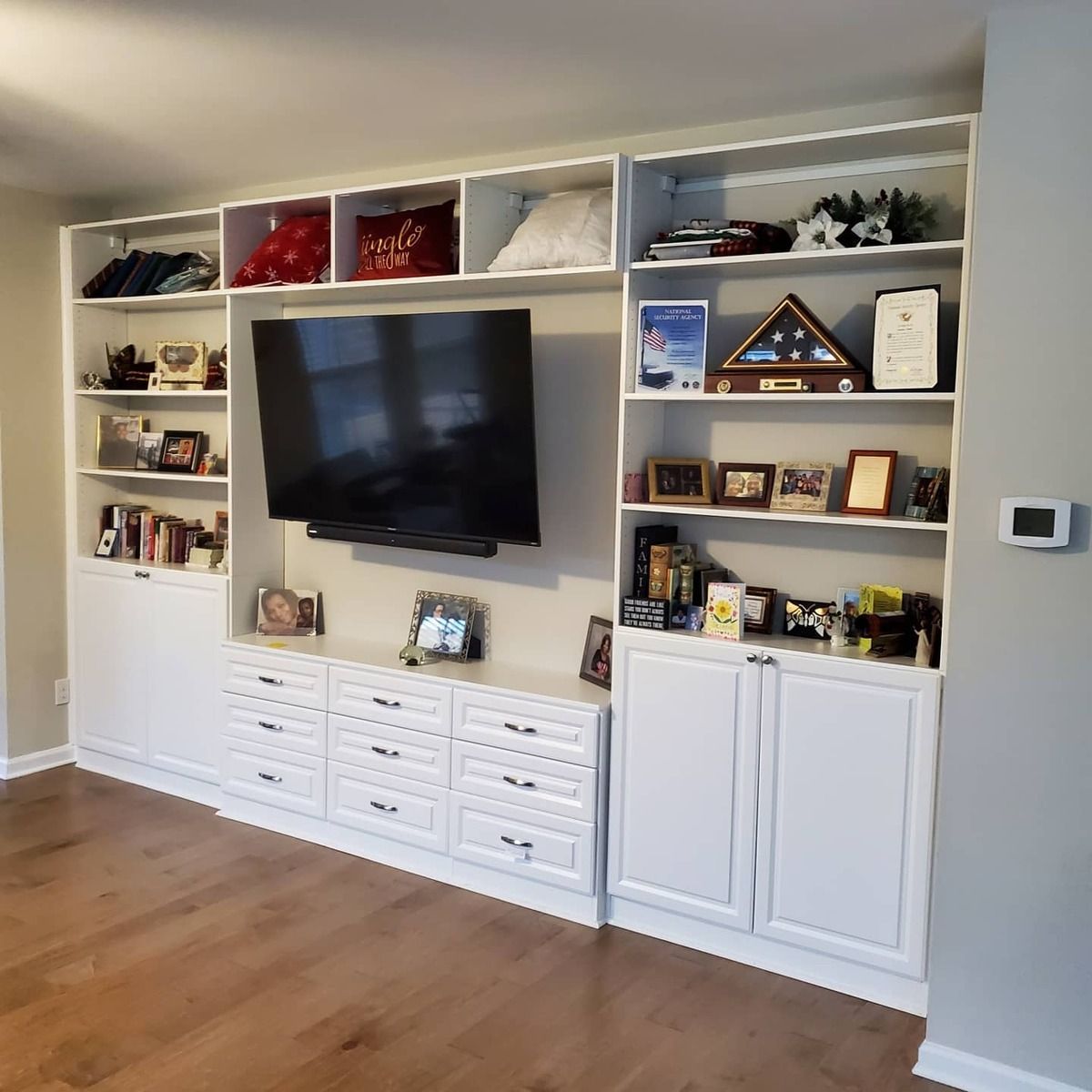 White built-in entertainment center with a TV, shelves, drawers, and cabinets filled with various items in a room with hardwood floors.