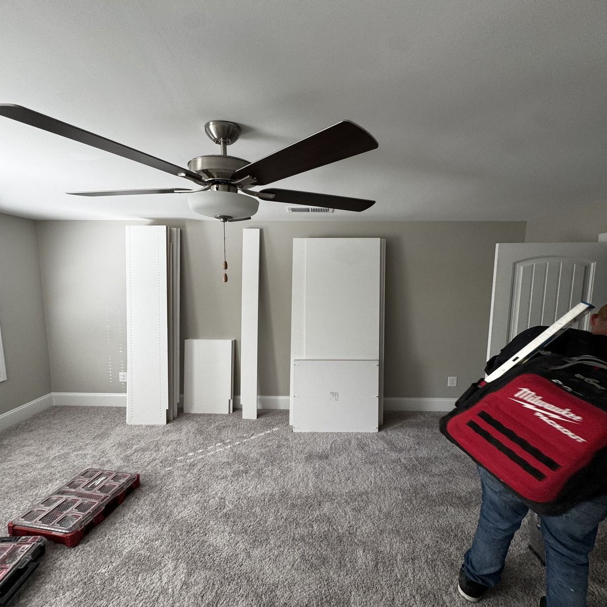A person carrying a tool bag is in a room with assembled furniture pieces against a wall and a ceiling fan.
