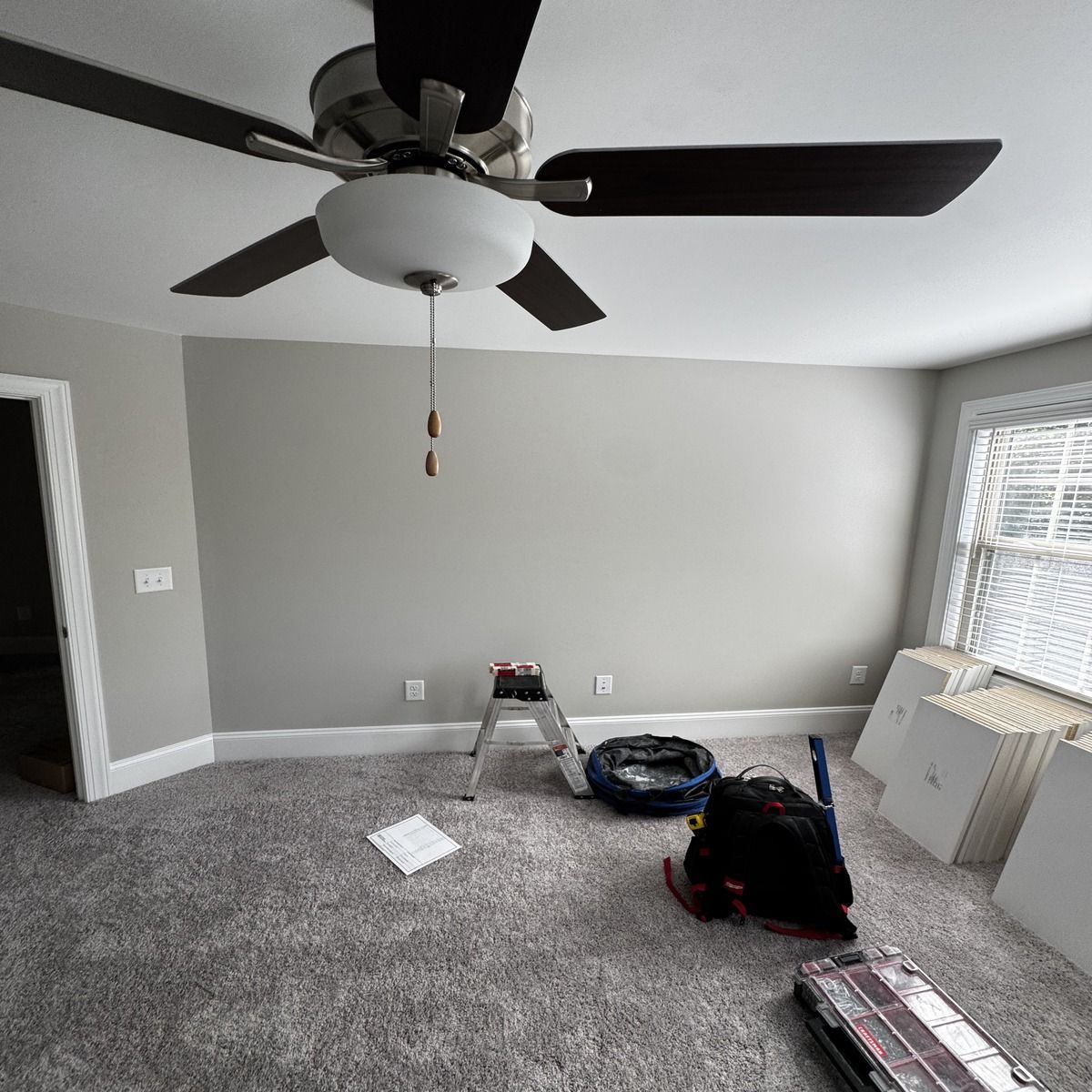 A room with a ceiling fan, grey walls and carpet, and a small stepladder. There's a person working on the floor.