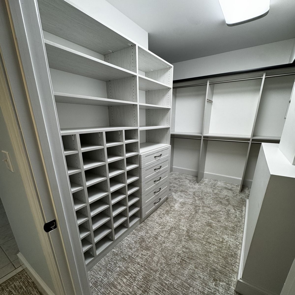 A walk-in closet with custom white shelving, drawers, and a shoe rack. The carpet is beige.