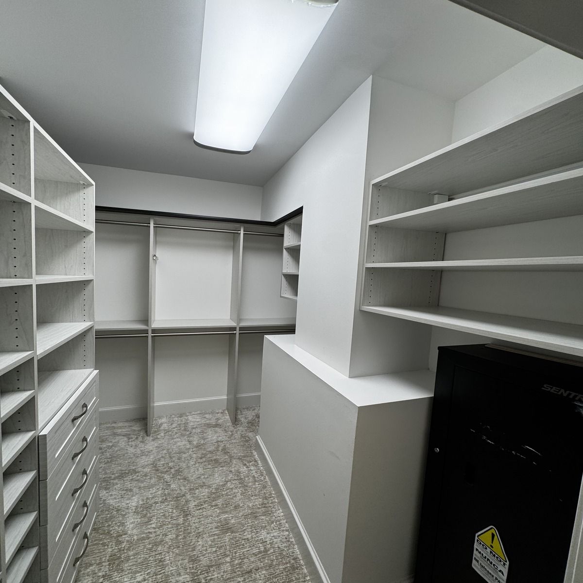Empty walk-in closet with white shelves, drawers, and carpet. The closet has an open doorway and a light fixture.