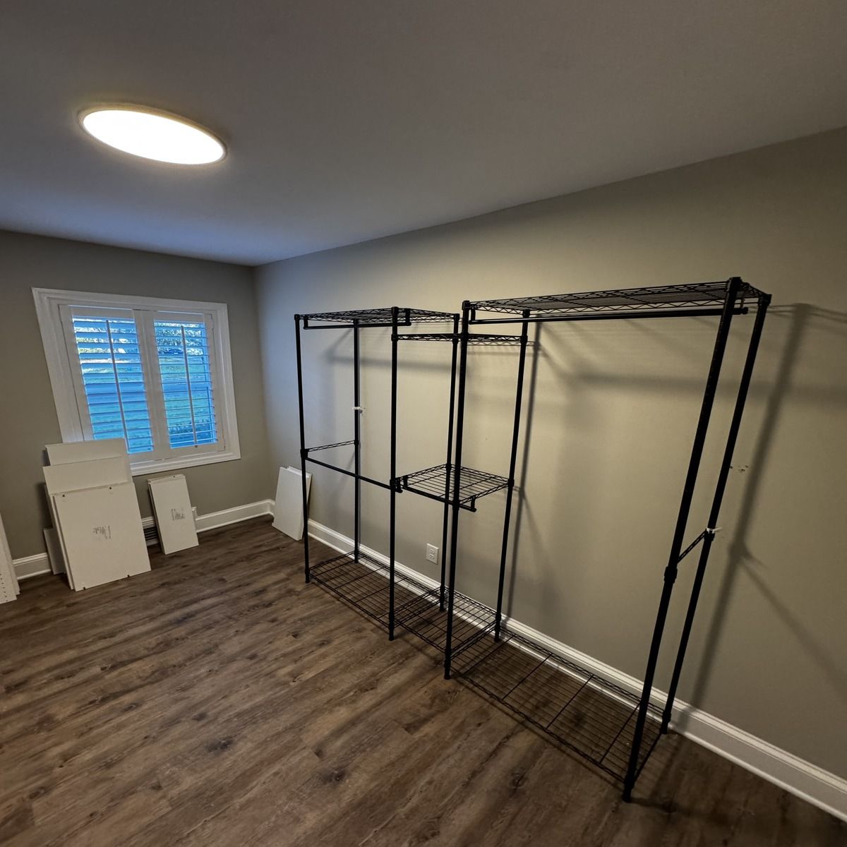 A room with dark wood flooring, a window with white shutters, and black wire shelving units against a tan wall.