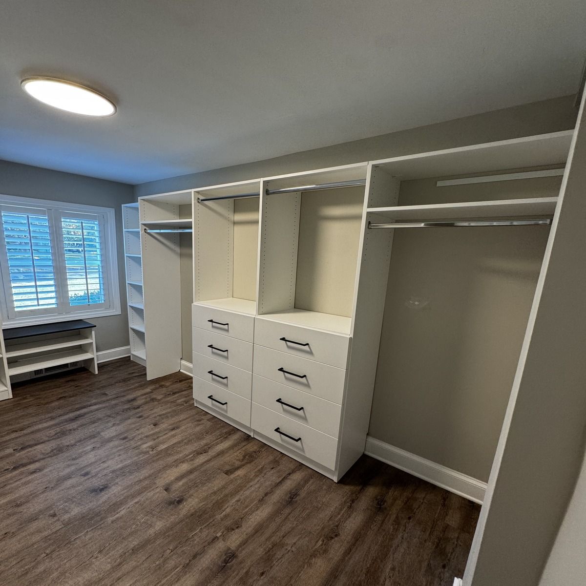 A bright, newly remodeled closet with white shelving and drawers, dark wood-look flooring, and a window in a light-gray walled room.