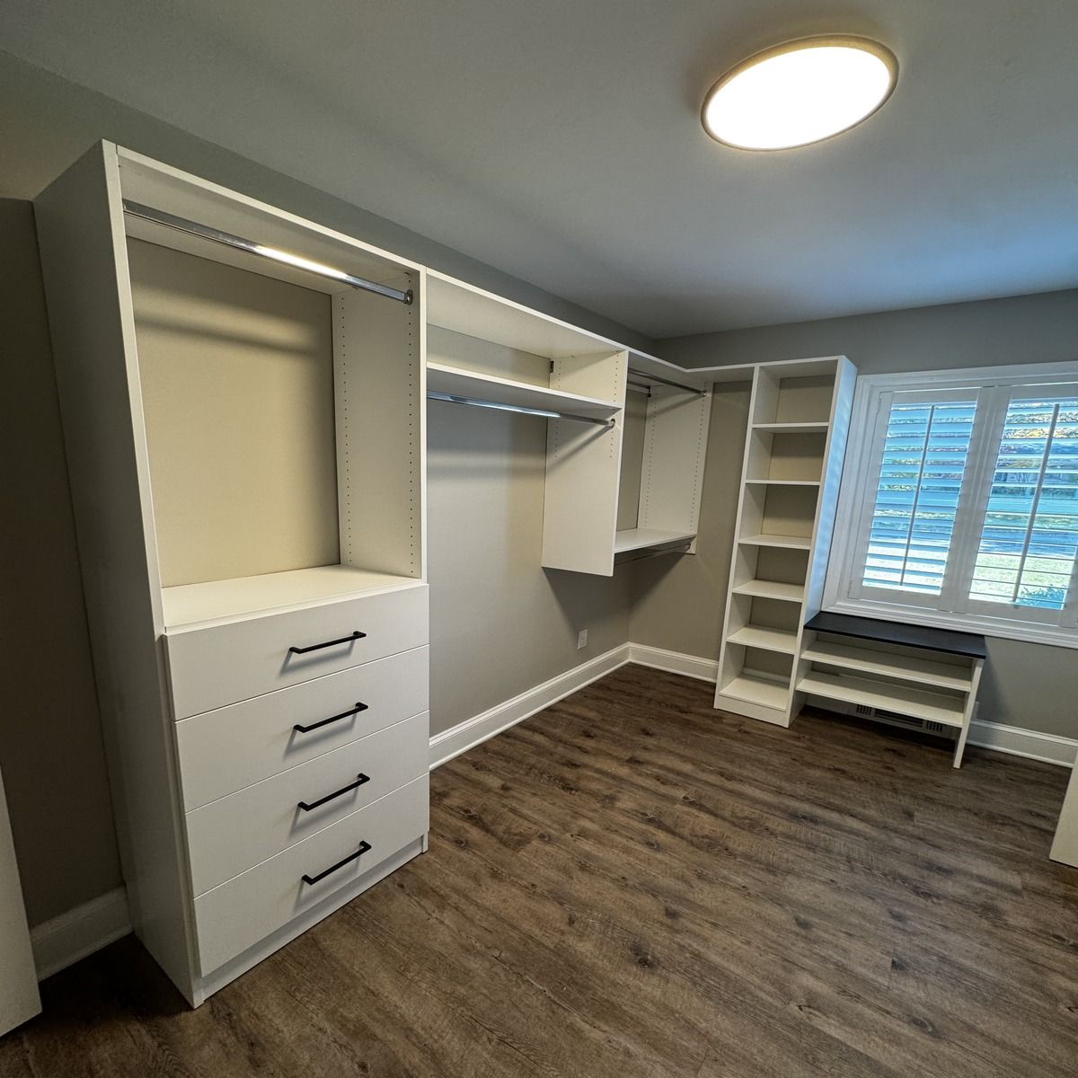 Walk-in closet with white built-in storage, drawers, and shelving. Dark wood-look floor and window with shutters.