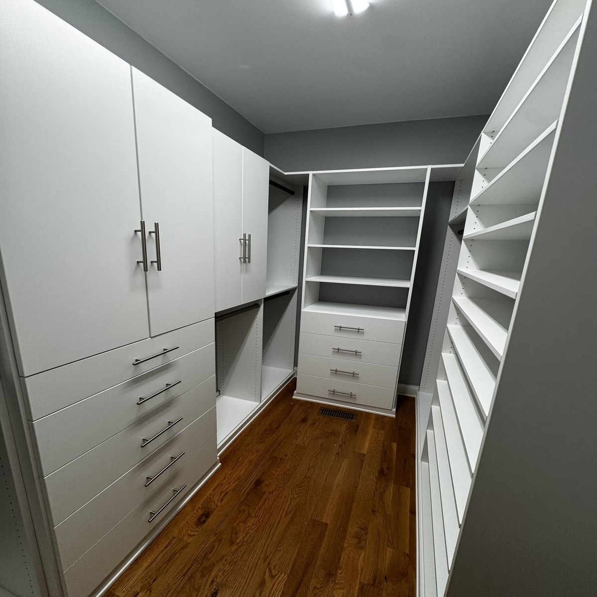 A modern, white walk-in closet with built-in shelving, drawers, and cabinets. It has wood flooring and gray walls.