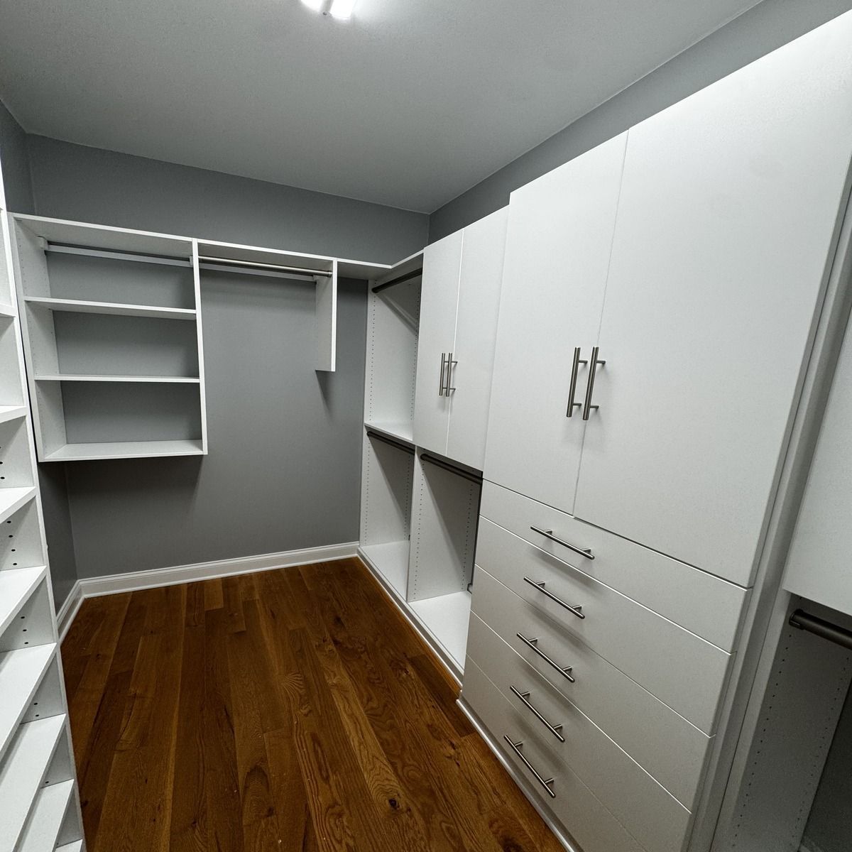 A walk-in closet with white shelving and cabinetry against gray walls and a dark wood floor.