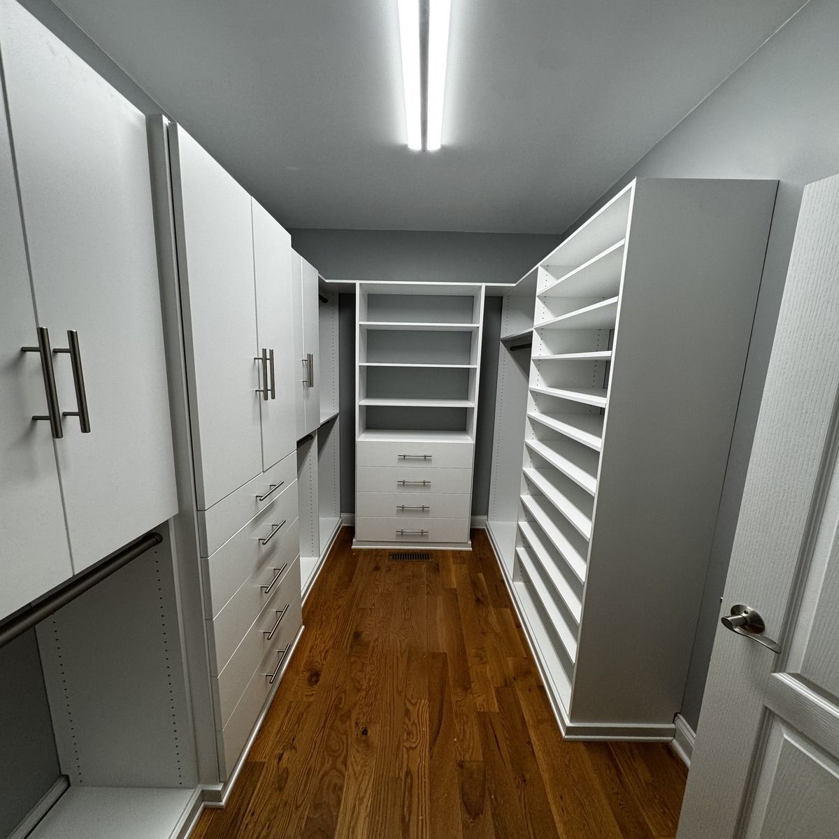 A well-lit, modern walk-in closet with white cabinetry, shelves, and drawers.  Dark wood flooring contrasts with the bright walls and fixtures.