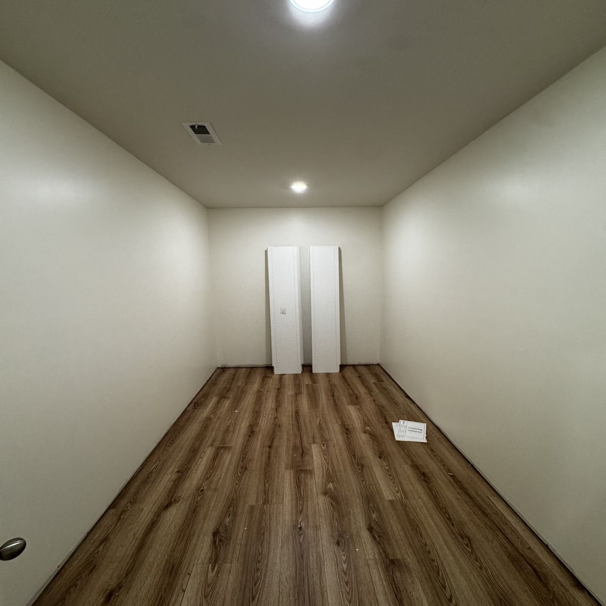 Empty, narrow room with light brown wood-look flooring and white walls. Two tall, white door panels stand against the back wall.