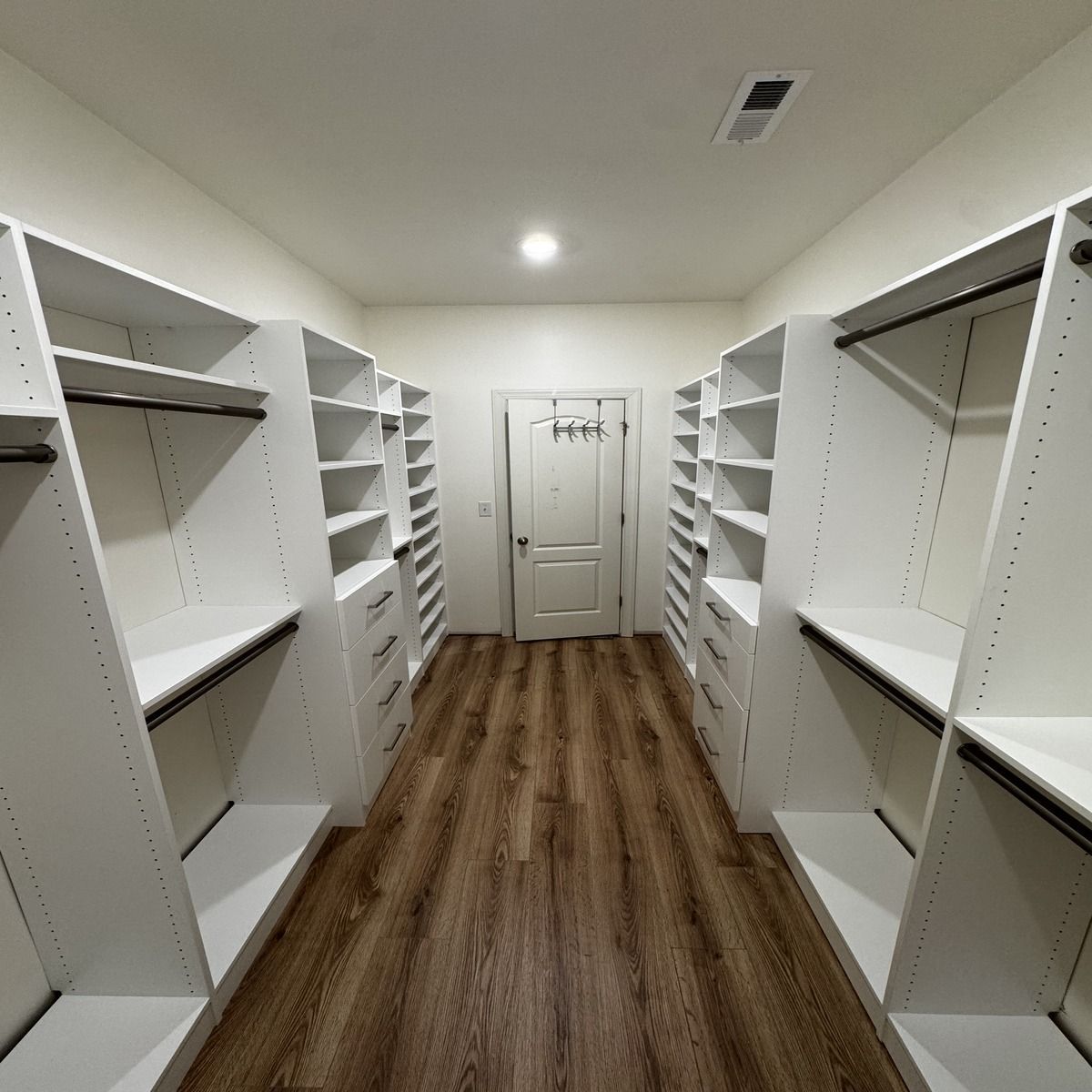 A spacious walk-in closet with white shelving and drawers on both sides, a light brown floor, and a door at the end.