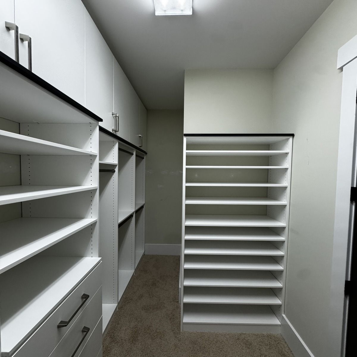 White walk-in closet with shelves and drawers, black trim, and a shoe rack. Beige carpet, neutral walls, and overhead lighting.