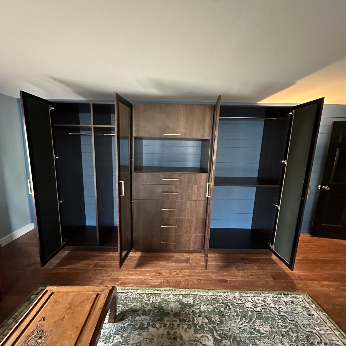 A large, custom-built closet with open doors reveals clothing racks and shelves. The dark wood structure sits on a hardwood floor with a rug.